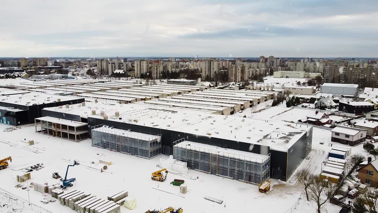 Logistics building under construction with Kaunas city residential district behind during snowfall, aerial ascend view