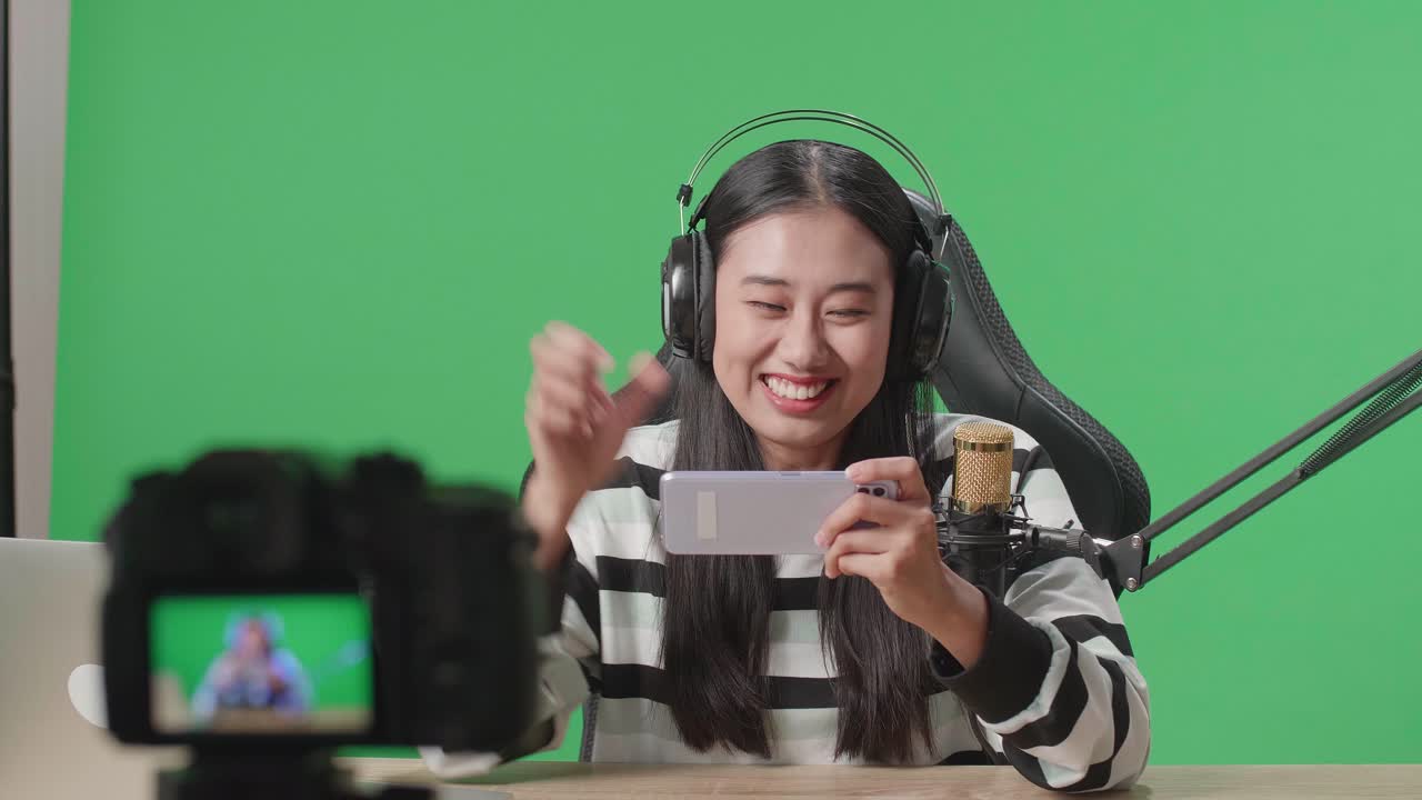 Gamer asiatica con cuffie e computer che festeggia la vittoria a un gioco per cellulare mentre riprende un video con la fotocamera su green screen
