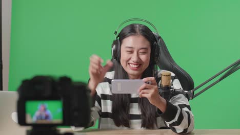 asian woman gamer with headphone and computer celebrating winning mobile phone game while shooting video by camera on green screen