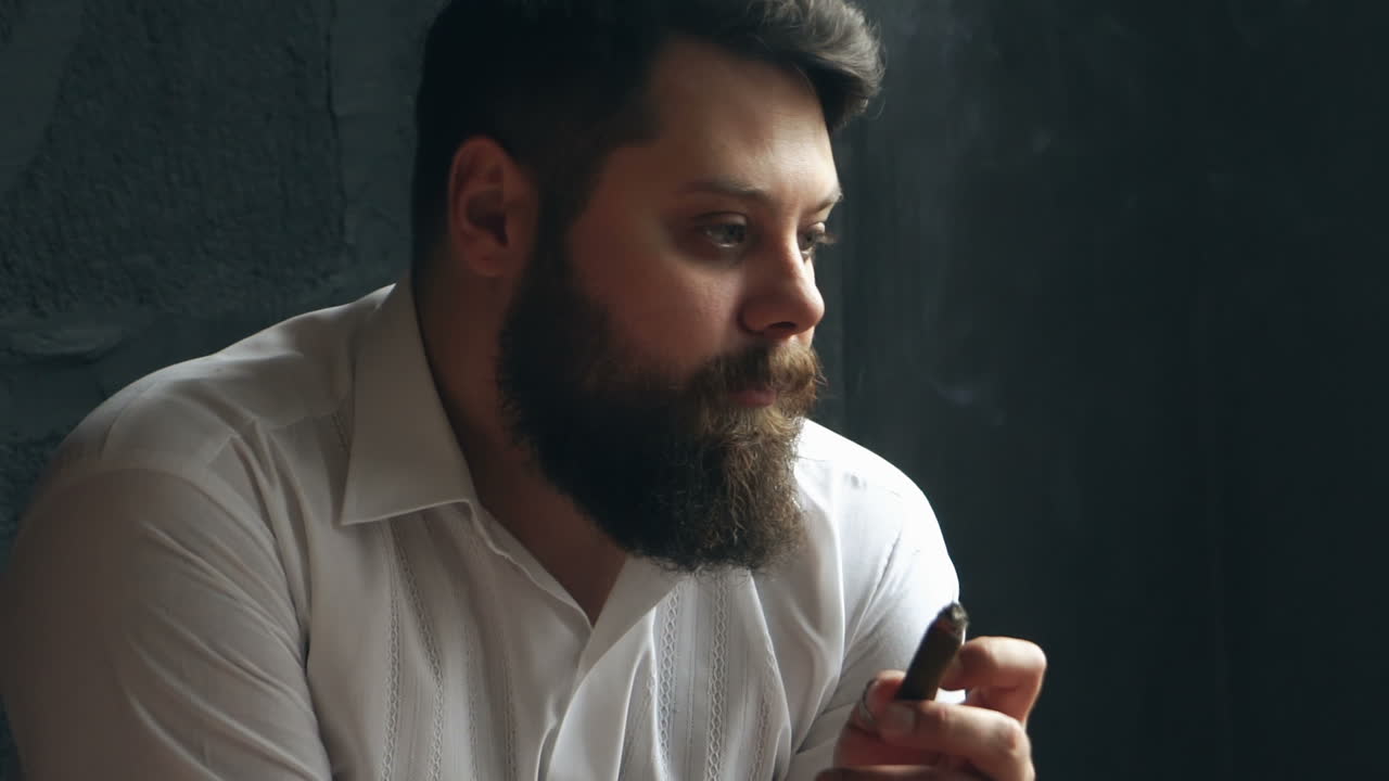 Uomo serio e rude in studio. Hipster caucasico serio e rude seduto e che fuma un sigaro.