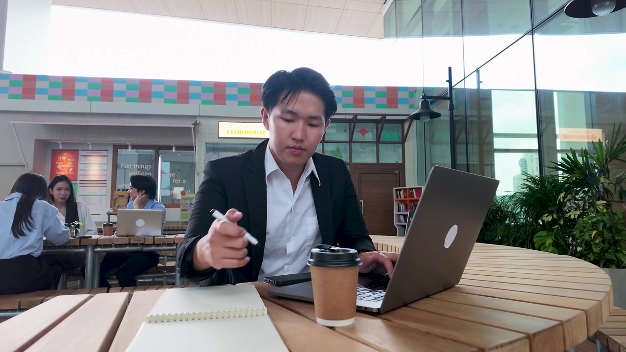 タイのバンコクの屋外カフェで、スーツを着たプロフェッショナルな男性がノートパソコンを使いメモを取る