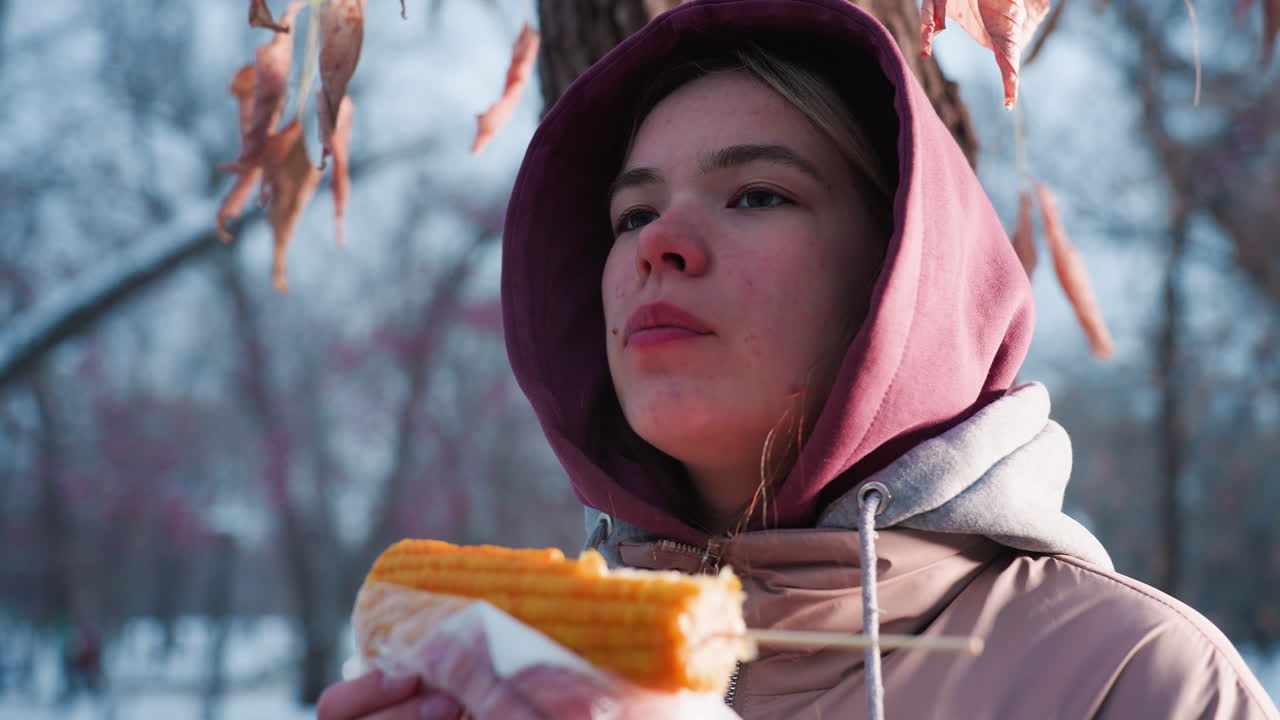 마른 잎이 부드럽게 흔들리는 야외 공원에서 옥수수를 먹는 여성, 겨울에 간식을 즐기고, 배경에 부드러운 빛 반사, 조용한 공원 분위기, 가운 바람과 함께 겨울 장면