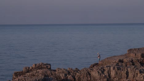 Hombre-Con-Sombrero-Pescando-En-Una-Roca-Junto-Al-Hermoso-Mar,-Hora-Dorada,-Espacio-De-Copias
