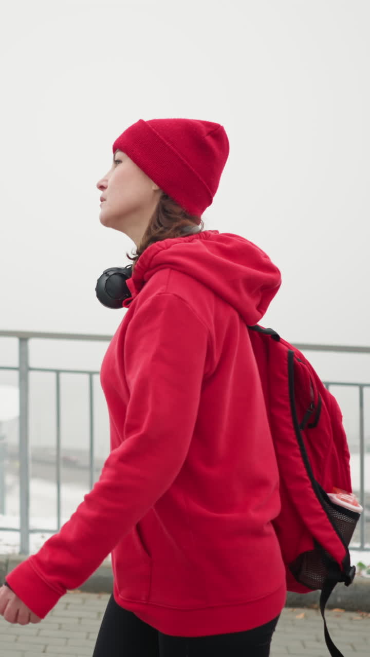 Side view of woman wearing red beanie and jacket walking along iron railing with headphones around neck and backpack, snowy landscape and blurred bridge and city in foggy atmosphere