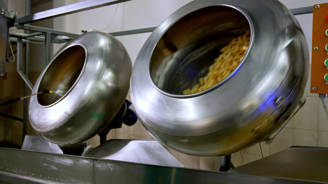 Two metal rotating tanks filled with sweet snacks. Corn sticks being covered with sugar syrup from a special disperse device.