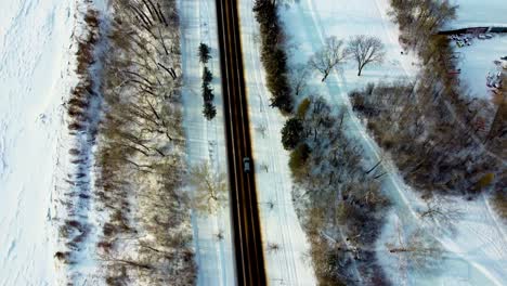 aerial birds eye view fly down winter road next snow covered river, empty polished skating track and rink on a sunny reflective afternoon following grey suv down 2 two lanes quiet roadway park 2-3