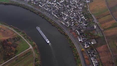 Cargo-ship-transporting-shipment-upstream-passing-idyllic-Bremm-town-on-Moselle-river,-aerial
