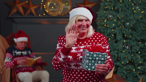 Senior-grandmother-in-Christmas-sweater-smiling-friendly-at-camera-and-waving-hands-gesturing-hello