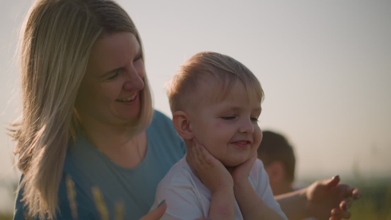 A loving mother in a blue dress enjoys a tender moment with her young son, holding his hands and kissing him with joy while sitting on the grass. A young boy, his older brother, is in the background