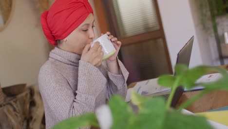 Video-of-biracial-woman-in-hijab-sitting-at-desk-at-home-working-on-laptop,-drinking-coffee
