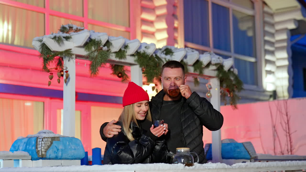 Loving Caucasian couple enjoying tea and each other's company outdoors in winter. Happy man and woman stand at the porch of the house talking, touching noses and smiling.