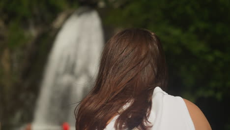 woman with messy brown hair against blurry waterfall at hike closeup. tourist observes heavy water flow falling from steep cliff on summer vacation