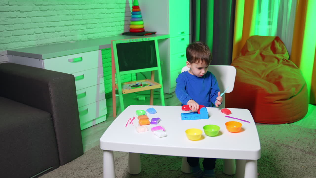 Child boy in blue sweater sits at desk using toy molding machine. Focused toddler plays with plasticine. High angle view.