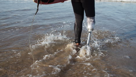 back view of a male surfer with bionic leg in wetsuit entering into ocean with surfboard under arm