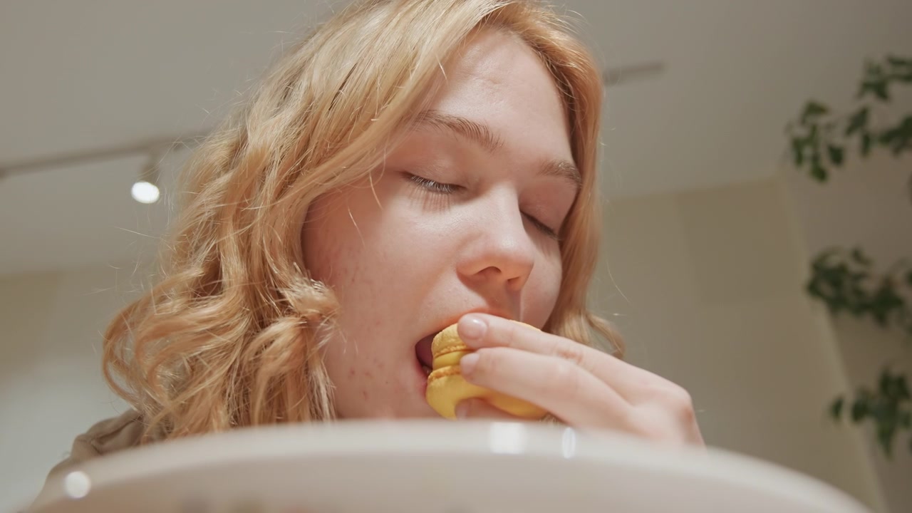 Close up of young woman eating yellow macarons indoors with soft lighting. Woman holds and takes bites from pastries focusing on her actions as woman enjoys them. scene features warm indoor