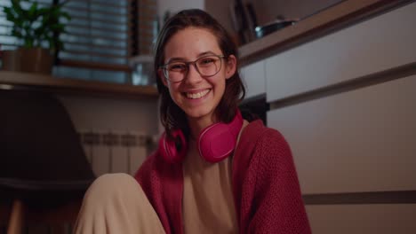 Portrait-of-a-happy-young-brunette-girl-in-glasses-red-wireless-headphones-and-a-sweater-who-is-sitting-and-posing-on-the-floor-in-the-kitchen-in-a-modern-apartment-in-the-evening