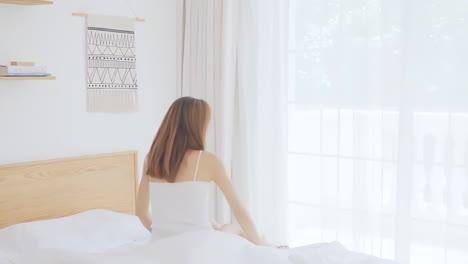 피자마를 입고 일어나는 아름다운 소녀의 실제, 큰 창문 근처에 앉아있는 침대에서 깨어나는, 완전히 휴식을 취하고, 아침에 신선한 공기와 빛을 얻기 위해 커튼을 펴는.
