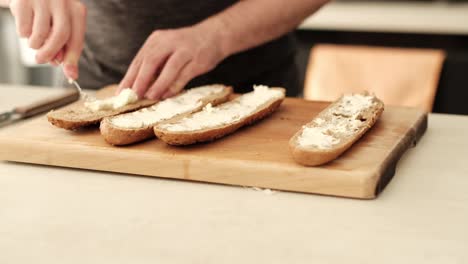 hombre caucásico extendiendo queso cremoso en una baguete de harina marrón en la encimera de la cocina