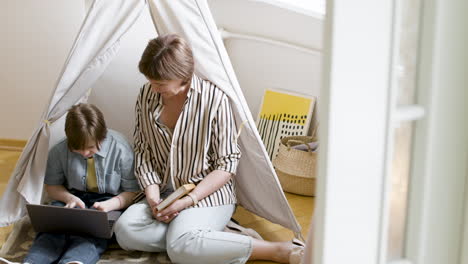 Young-girl-using-laptop-and-kissing-her-mother,-who-is-next-to-her-holding-a-book