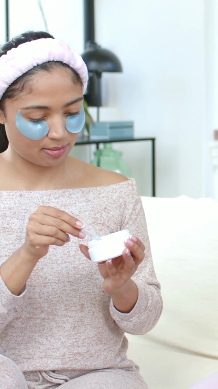 Vertical video: Starting woman in pink headband, blue gels scooping cream on sofa, with jar, scoop