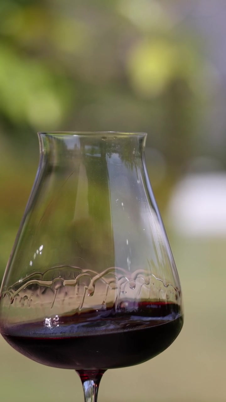 Primo piano verticale di vino rosso che volteggia in un bicchiere su uno sfondo di giardino delicatamente sfocato.