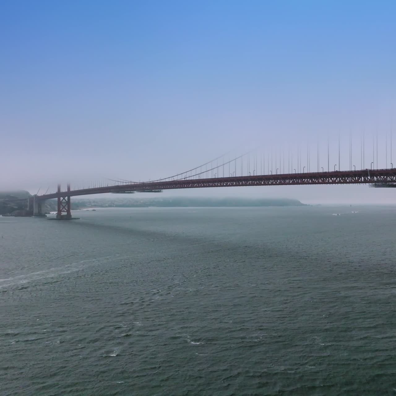 Beautiful Golden Gate Bridge covered with thick white fog. Boats sailing by the straight under the metal construction. Side view