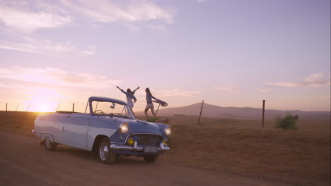 Amigas-Bailando-Al-Atardecer-En-Un-Viaje-Por-Carretera-Con-Un-Coche-Antiguo