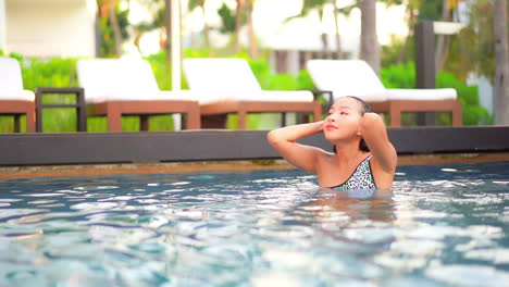 a sexy young woman emerges from the water of a resort swimming pool pushing her wet hair away from her face