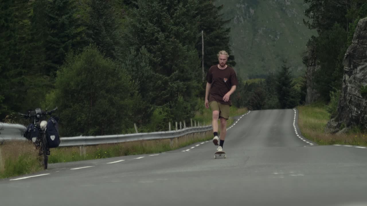 Giovane skateboarder maschio caucasico che sale con lo skateboard verso la telecamera su una strada desolata nel mezzo del nulla in Norvegia, Scandinavia