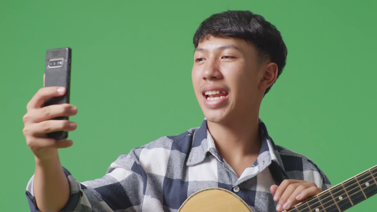 녹색 화면 배경에 서 있는 동안 기타를 흔들고 스마트폰으로 비디오 통화를 하는 아시아 십대의 클로즈업
