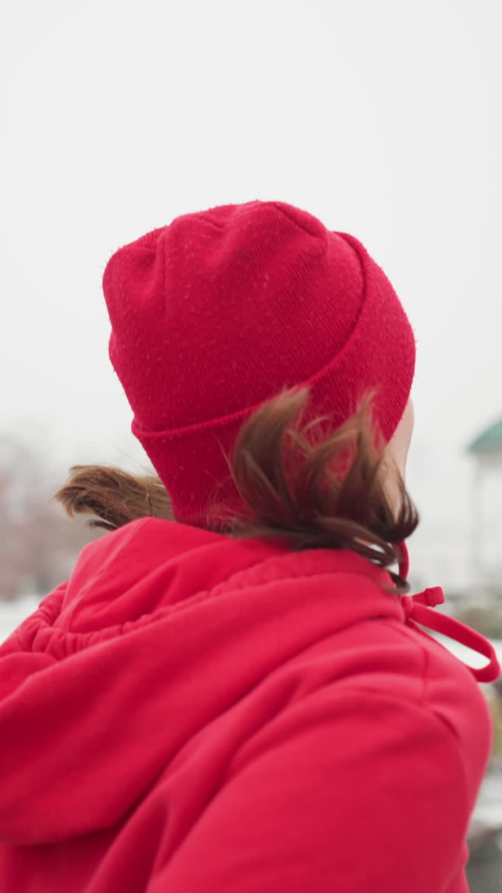 lady jogging along snowy pathway wearing red beanie and jacket hair fluttering in cold breeze surrounded by benches colorful bushes gazebo and serene foggy winter scenery conveying fitness motion