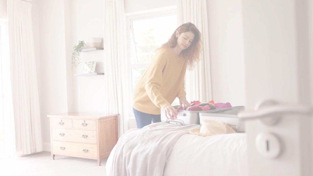 Người phụ nữ mở vali và sắp xếp đồ đạc trên giường cạnh cửa ra vào, di chuyển đôi giày màu hồng, chuẩn bị cho chuyến đi.