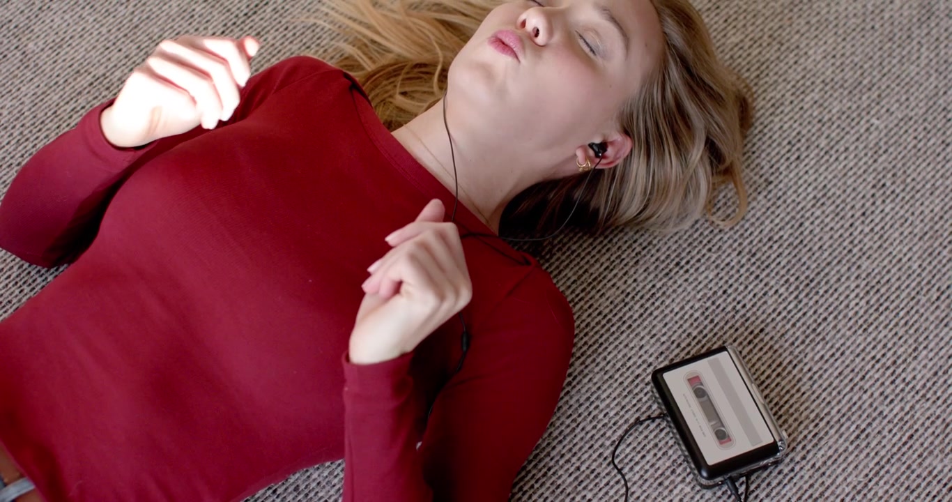Woman in her 20s lying on carpet in burgundy top, cassette player playing, moving hands, mouthing