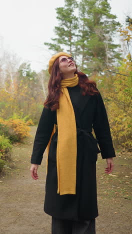 mujer elegante en abrigo negro con boina amarilla y bufanda se encuentra en el parque de otoño, mirando a su alrededor y girando mientras toca su cabello rojo, colores de otoño vibrantes la rodean en un entorno al aire libre pintoresco