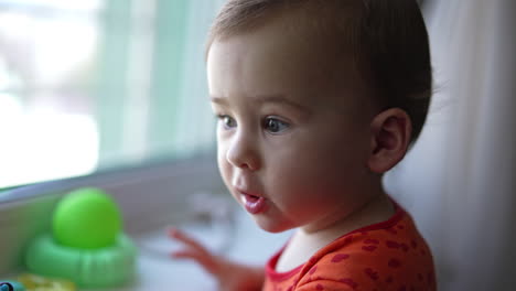 lovely caucasian kid standing at the window-sill. baby makes a wry face moving forward to the camera. close up. blurred backdrop.