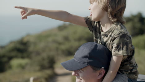 close up of father carrying son on shoulders on hiking adventure while boy pointing at something in distance