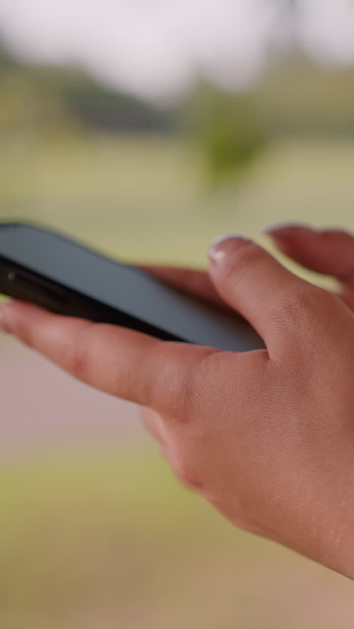 primer plano de la mano de un individuo escribiendo en un teléfono inteligente con enfoque en las manos, en contra de un fondo verde naturalmente borroso con figuras poco claras en la distancia