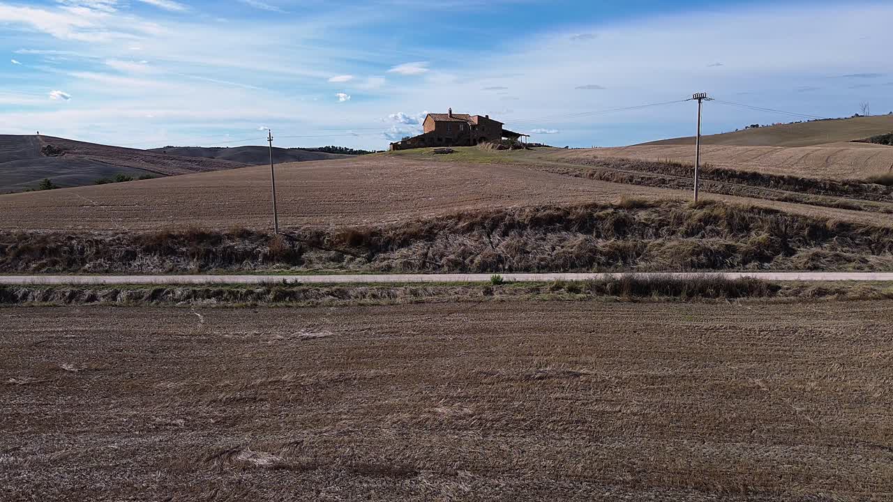 Rustic farmhouse nestled among Tuscany's rolling hills, surrounded by golden fields and dirt roads under a blue sky.