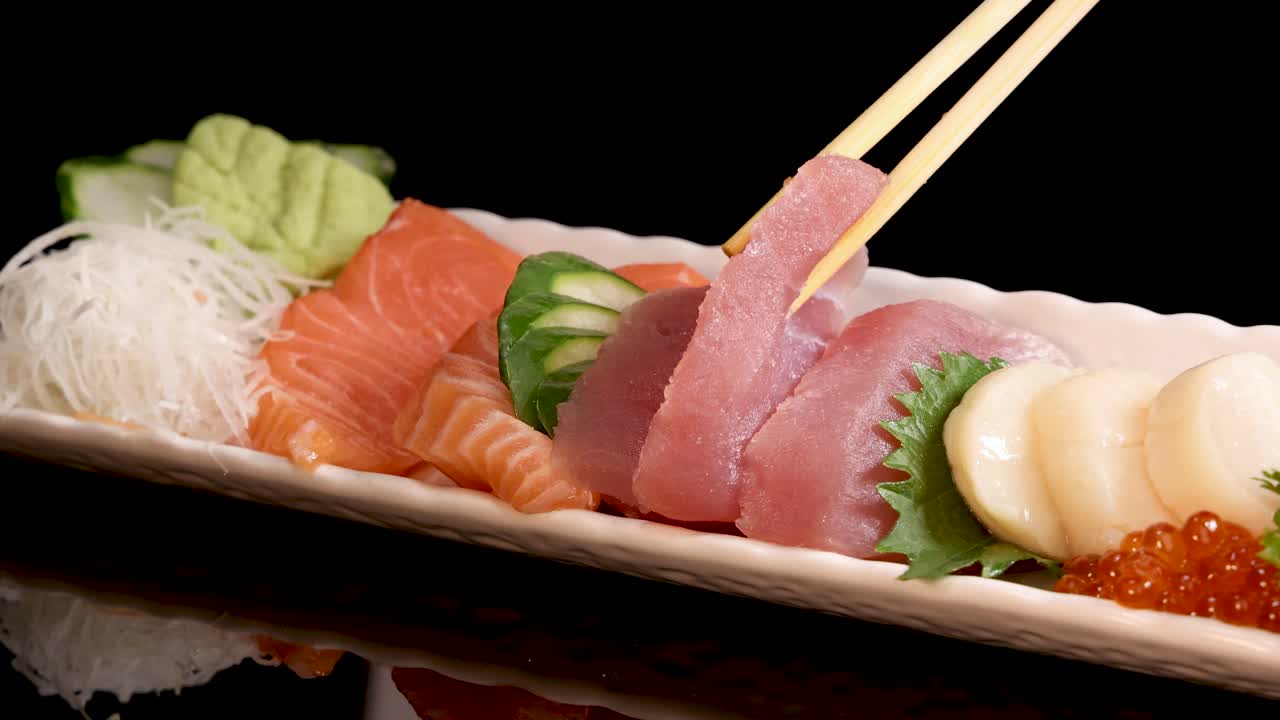 Chopsticks delicately pick up a slice of tuna sashimi from a Japanese sushi set, presented on a black background with dramatic, focused lighting