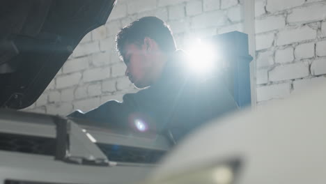 car engineer in blue uniform works on car engine in auto workshop with bright light shining from background, mechanic leans over vehicle, concentrating on detailed repairs in professional setting