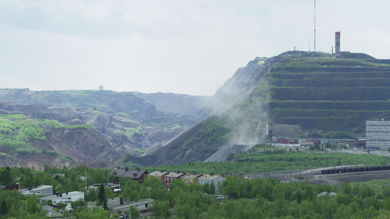 Medium shot of iron ore mine run by Luossavaara-Kiirunavaara AB LKAB in Kiruna, Norrbotten, Lapland, Sweden. Dust rising from the rocky talus slope at the bottom of the mine benches. Summer, day