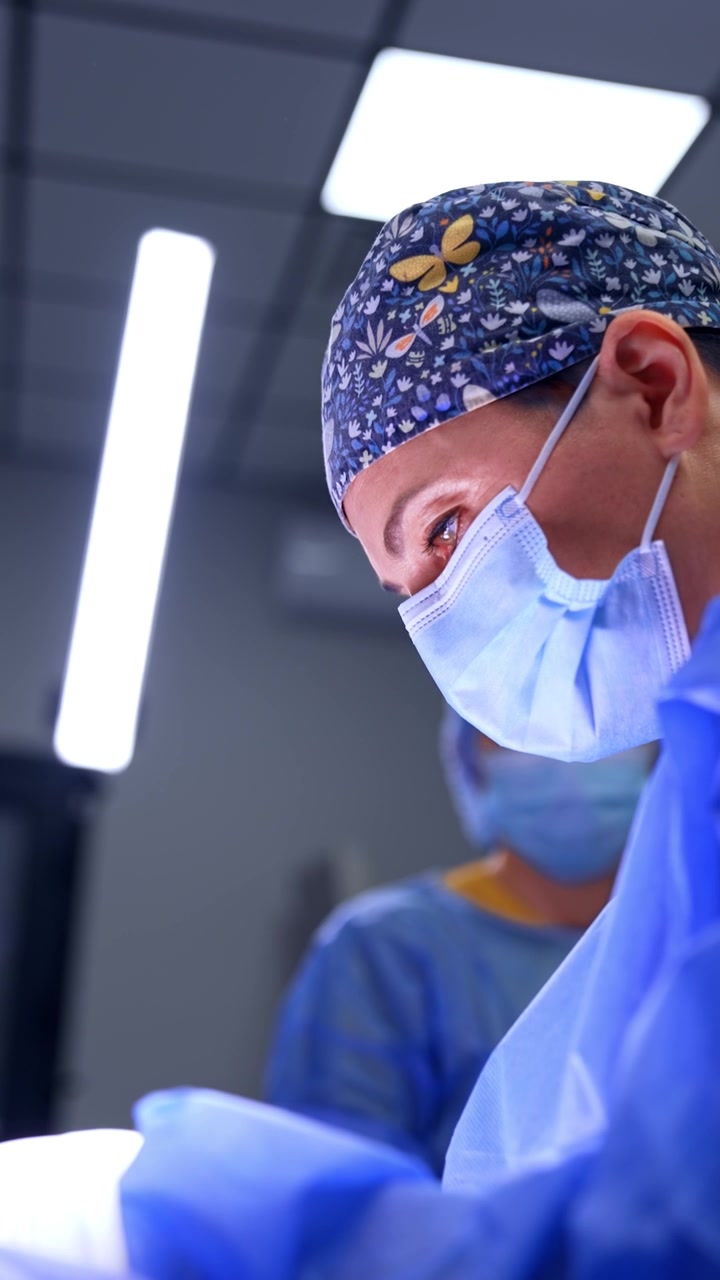 Medico professionista con cuffia a fantasia floreale e mascherina è concentrato sull'operazione. Vista dal basso del chirurgo che lavora in sala operatoria. Video verticale