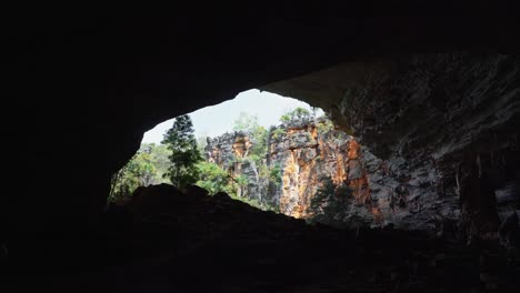 Tiro-Inclinado-Desde-El-Interior-De-La-Cueva-Lapa-Doce-Mirando-Hacia-Afuera,-Revelando-Una-Enorme-Entrada-A-La-Cueva-Con-Coloridos-Acantilados-Y-Follaje-Selvático-En-El-Parque-Nacional-Chapada-Diamantina-En-Bahia,-Noreste-De-Brasil