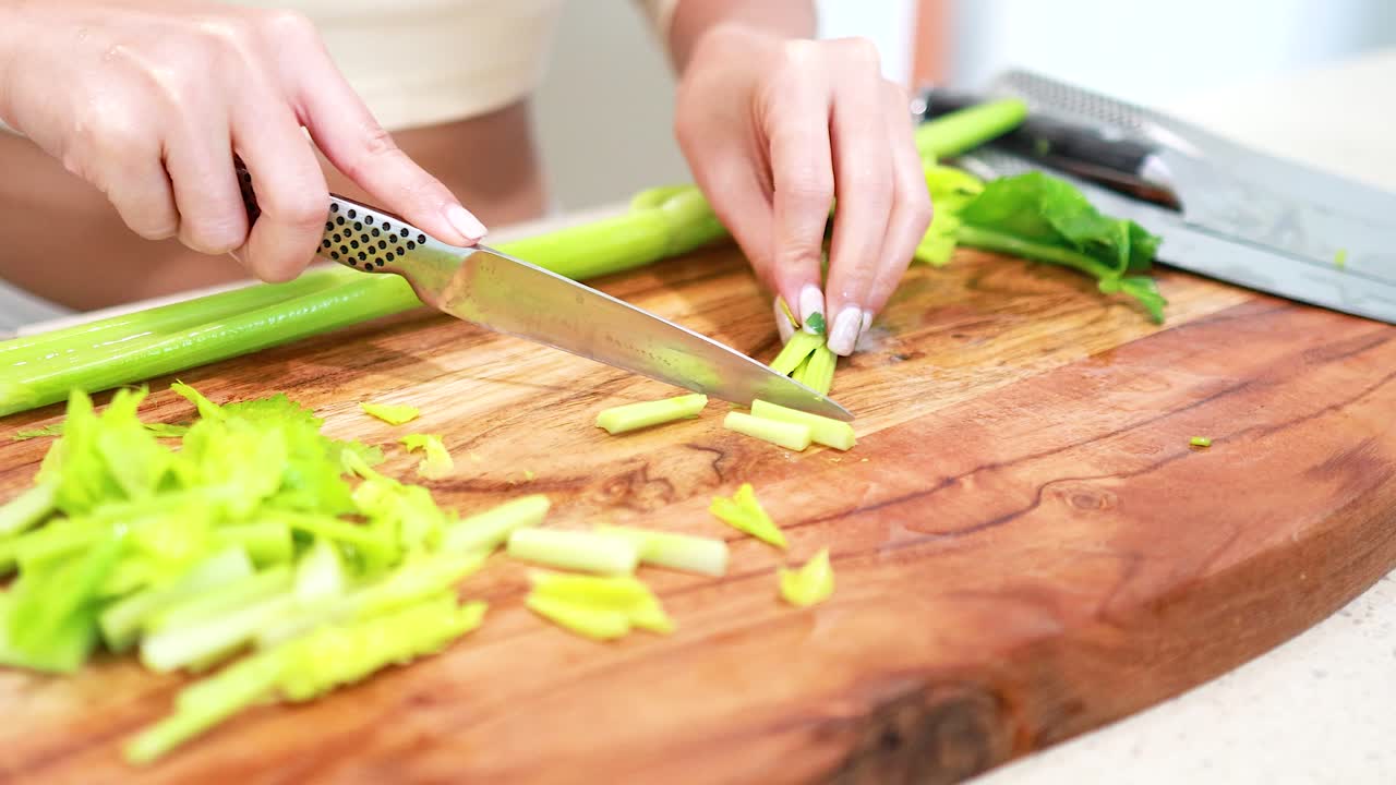 Hands slicing celery on a wooden board in a bright kitchen. Close-up view with natural lighting and detailed focus