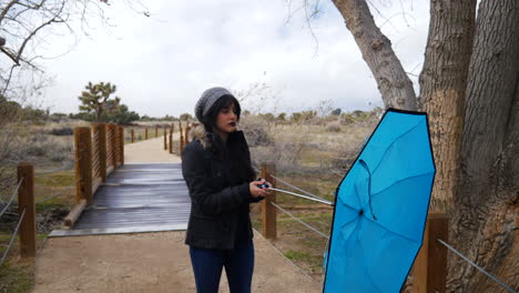 una mujer abre un paraguas azul en una tormenta de lluvia que se vuelve del revés mientras frunce el ceño decepcionada por el mal tiempo