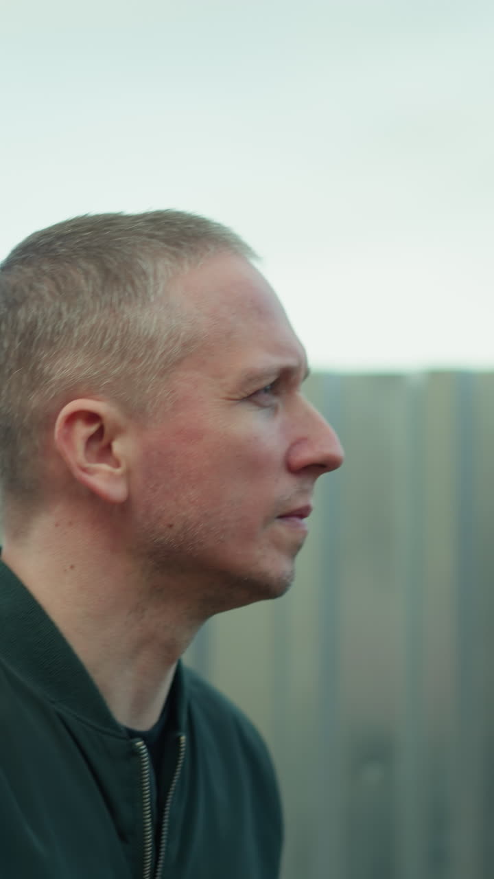 A close view of a man in a green jacket looking up thoughtfully while standing next to a corrugated metal fence