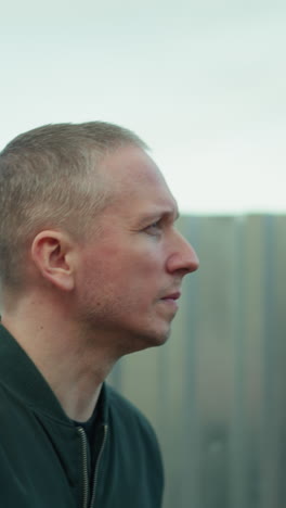 a close view of a man in a green jacket looking up thoughtfully while standing next to a corrugated metal fence