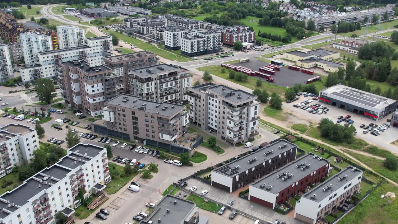 Aerial view of mixed residential blocks with mid-rise apartments, townhouses, parked cars, green courtyards, and nearby commercial areas on the edge of a growing suburban district