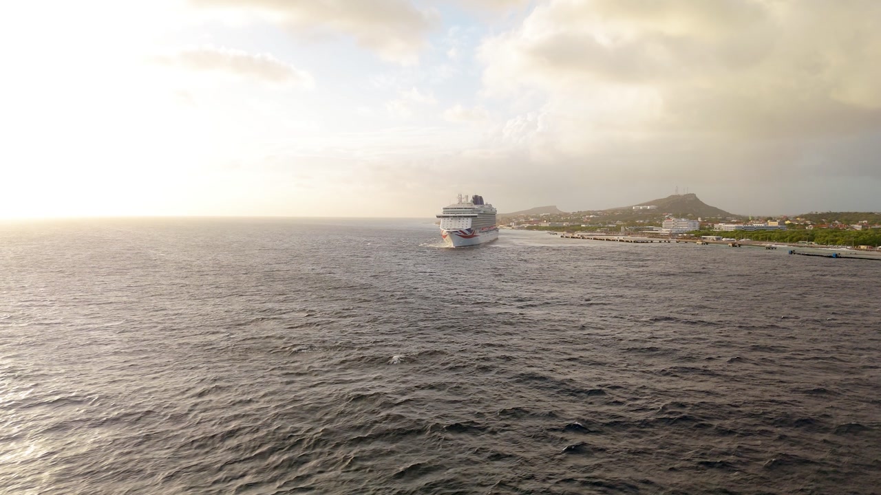 Wide drone shot of a cruise ship leaving port during sunset with glowing sky over Curacao waters.