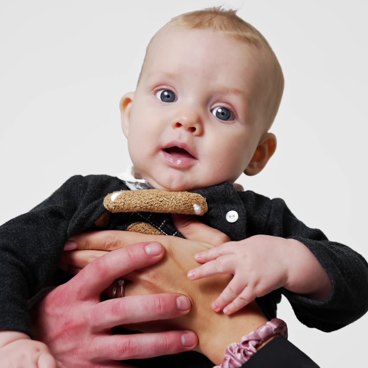Male and female hands hold beautiful smiling Caucasian infant. Mother and father kiss their lovely son on the head. White backdrop